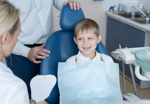 Portrait of smiling  little boy sitting in dental chair  listening to female dentist holding tooth model explaining oral hygiene rules with father at his side